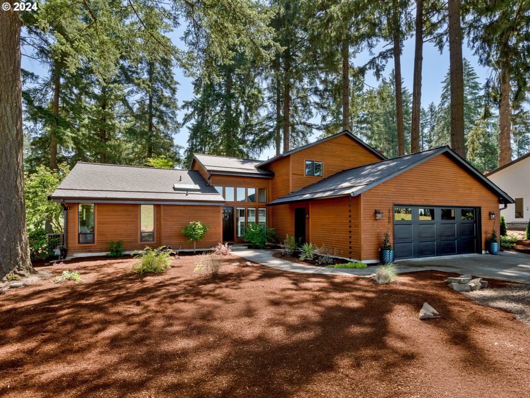 Contemporary wood house with a dark roof and two-car garage, surrounded by tall trees and landscaped yard.